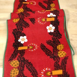 Puede incluir: Caminero de mesa rojo con borde verde que presenta un patrón repetido de ramas de pino, piñas, velas y flores blancas.