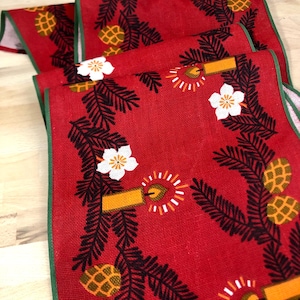 May include: Red table runner with a green border featuring a repeating pattern of pine branches, pine cones, white flowers, and yellow candles.