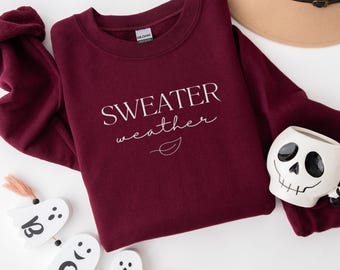 May include: A burgundy sweatshirt with the words "SWEATER weather" in white script. A tan hat with a black band and a skull-shaped mug are also visible. Decorative wooden ghosts with tassels are in the foreground.