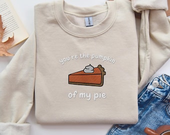 Può includere: Felpa beige con la frase ricamata "you're the pumpkin of my pie" e una grafica a forma di fetta di torta di zucca. La felpa è piegata, con un libro, jeans e una tazza sullo sfondo.