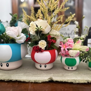 May include: A set of four mushroom-shaped planters in various sizes and colors. The planters are painted in blue, red, and green with white accents and filled with colorful flowers and greenery. Each planter has two black eyes.