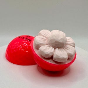 Könnte beinhalten: Ein rosa Golfball mit dem aufgedruckten Wort "RESIS" ist aufgespalten, um ein weißes, blumenförmiges Objekt im Inneren zu enthüllen.
