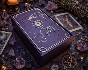 May include: A dark purple wooden box with a gold-colored design on the lid, featuring hands reaching for a celestial symbol. The box is surrounded by crystals, tarot cards, and a lit candle, creating a mystical aesthetic.