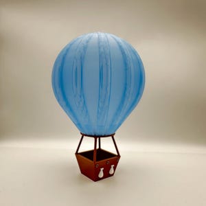 May include: A decorative hot air balloon with a light blue balloon and a brown basket. The balloon has vertical lines and a textured appearance. The basket has two white decorative elements.