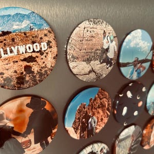 Pode incluir: Uma coleção de ímãs redondos com várias imagens, incluindo uma paisagem com a palavra "HOLLYWOOD" em letras brancas, uma cena do deserto, uma cordilheira, um céu com nuvens, uma pessoa em um barco, um fantasma de desenho animado, um horizonte da cidade e uma pessoa em uma floresta.