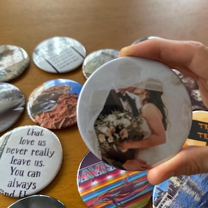 May include: A collection of round buttons with various designs. One button features a wedding photo of a couple kissing. Other buttons display text, landscapes, and a dolphin graphic. The buttons are on a wooden surface.