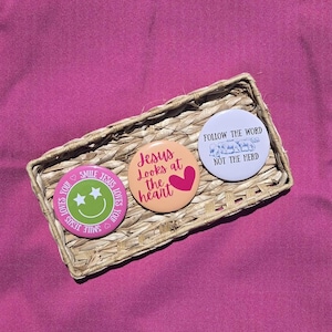 May include: Three round pinback buttons with religious themes rest in a woven basket on a pink surface. One button has a green smiley face with stars and the text "SMILE JESUS LOVES YOU". Another says "Jesus Looks at the heart" with a pink heart. The third says "FOLLOW THE WORD NOT THE HERD".