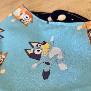 May include: Close-up of a light blue fabric with a repeating pattern of cartoon dogs. The dogs are in various poses, with orange, blue, and white coloring. The fabric is folded, revealing a dark blue interior with white polka dots.