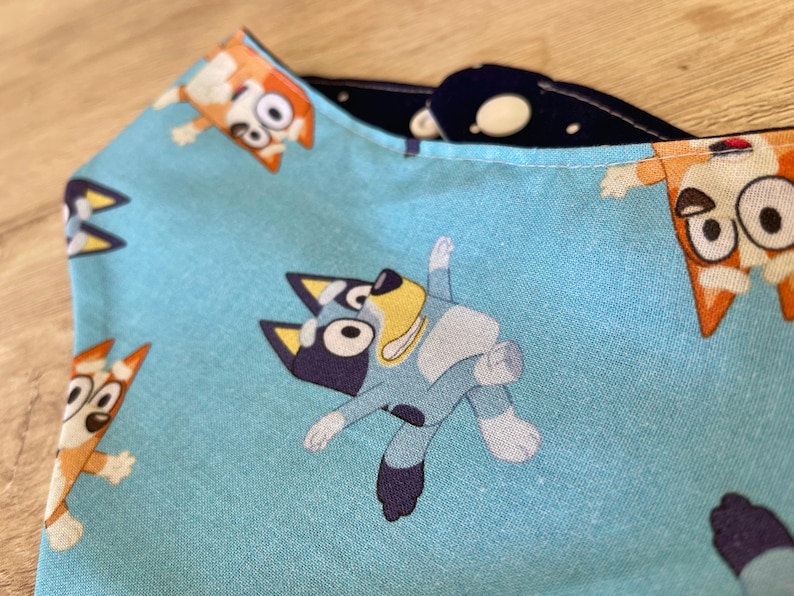 May include: Close-up of a light blue fabric with a repeating pattern of cartoon dogs. The dogs are in various poses, with orange, blue, and white coloring. The fabric is folded, revealing a dark blue interior with white polka dots.