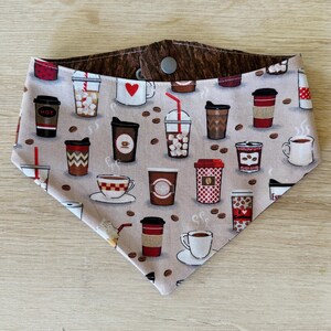 May include: A tan and brown dog bandana with a coffee theme. The bandana features various coffee cups, coffee beans, and coffee-related graphics. The bandana is made of fabric and has a snap closure.