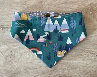 May include: A teal dog bandana with a camping-themed pattern. The design includes mountains, trees, tents, a rainbow, and a camper. The reverse side is gray with white polka dots, and it fastens with a single button.