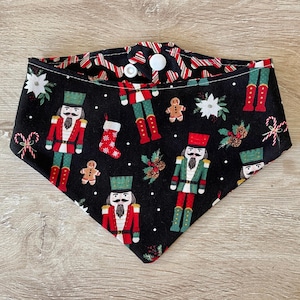 May include: Black bandana with a Christmas nutcracker pattern. The bandana features red, green, and white nutcrackers, candy canes, gingerbread men, and holly. The bandana has a white button closure.
