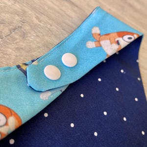 May include: A baby bib with a blue and orange cartoon character print on one side and a navy blue polka dot pattern on the other. The bib has two white snap closures.