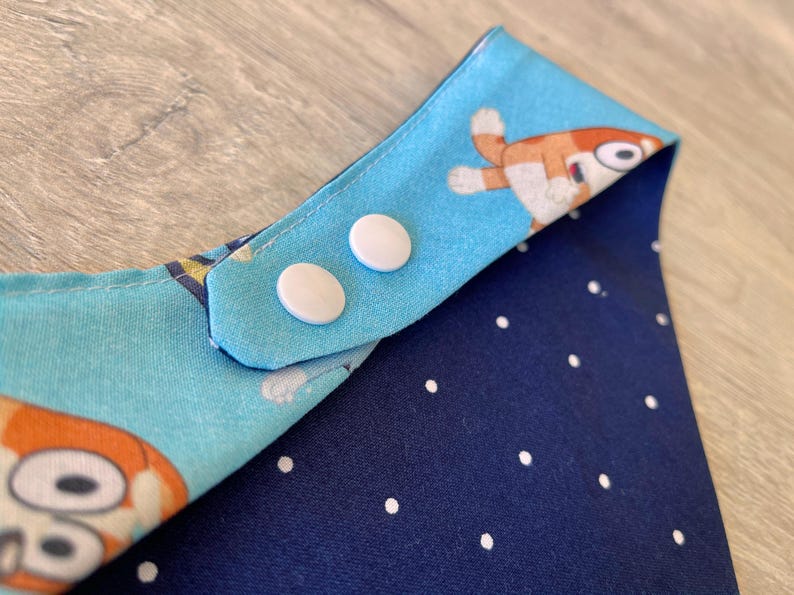 May include: A baby bib with a blue and orange cartoon character print on one side and a navy blue polka dot pattern on the other. The bib has two white snap closures.