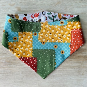 May include: A triangular pet bandana featuring a patchwork design of various floral and checkered patterns in shades of green, yellow, blue, and red. The fabric displays sunflowers and small white flowers, secured at the top with two white plastic snap buttons.