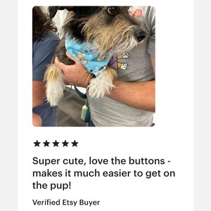May include: A small dog wearing a light blue bandana with a cartoon design. The dog is held by a person. The bandana has a button closure. The text "Super cute, love the buttons - makes it much easier to get on the pup!" is visible.
