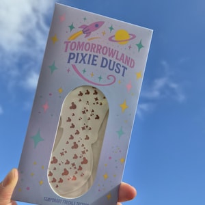 May include: A package of temporary freckle tattoos labelled "Tomorrowland Pixie Dust." The package is light blue with a rocket ship and planet graphic. The tattoos are white with brown Mickey Mouse head shapes and stars. The background is a bright blue sky.