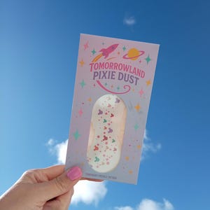 May include: A light purple card with the words "Tomorrowland Pixie Dust" and a rocket ship illustration. The card contains temporary freckle tattoos with colourful Mickey Mouse head designs. The background is a bright blue sky.