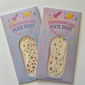 May include: Two packages of temporary freckle tattoos labelled "Tomorrowland Pixie Dust." The packages are light purple with a rocket ship and planet design. The tattoos are shaped like a silhouette with colourful dots.