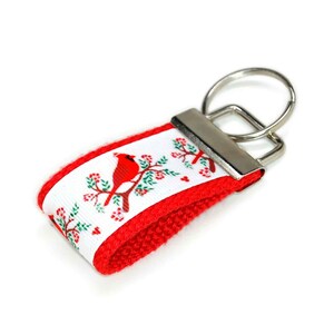 May include: Red cardinal bird key chain with a white background and red trim. The key chain has a silver key ring.