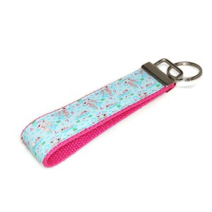 May include: A pink and blue key chain with a silver key ring. The blue fabric has a pattern of otters swimming in a pond.