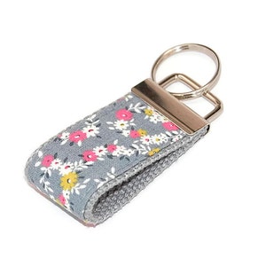 May include: A gray fabric keychain with a floral pattern in pink, white, and yellow. The keychain has a silver metal ring.