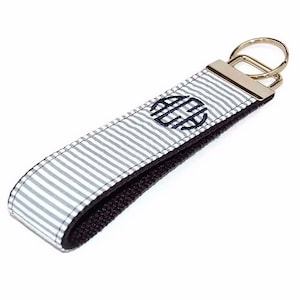 May include: A white and gray striped key chain with a black strap and a silver key ring. The key chain has a navy blue monogram in the center.