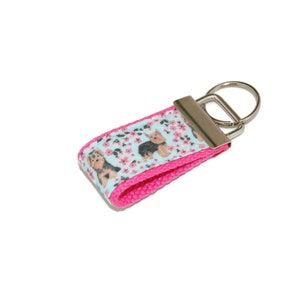 May include: A pink and blue key chain with a silver key ring. The key chain features a repeating pattern of Yorkshire Terriers and pink flowers on a light blue background.
