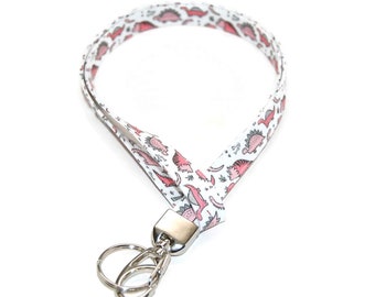May include: White lanyard with a repeating pattern of pink dinosaurs and green foliage. The lanyard has a silver-colored metal clasp and key ring. The fabric is folded over and sewn to create the lanyard.