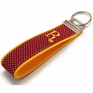 May include: A maroon and white polka dot keychain wrist strap with a yellow border and a gold embroidered letter "R". The keychain has a silver metal ring for keys.