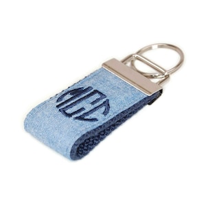 May include: A blue denim keychain with a navy blue monogram in a circle. The keychain has a silver metal ring.