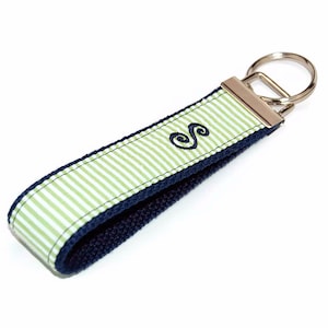 May include: A green and white striped key chain with a navy blue trim and a silver key ring. The key chain has a green embroidered letter "S" on it.