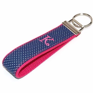 May include: A navy blue and pink key chain with white polka dots. The keychain has a silver key ring and the letter "K" embroidered in pink.