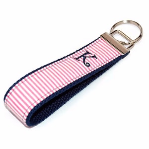 May include: A pink and white striped key chain with a navy blue trim and a silver key ring. The keychain has a navy blue embroidered letter "K" on it.