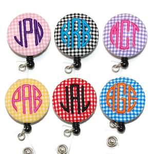 May include: Six different colored gingham fabric badge reels with a monogrammed initial in the center of each. The colors are pink, black and white, purple, yellow, red, and blue.