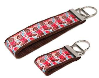 May include: Two brown keychains with silver key rings. The keychains are decorated with a pink and white ribbon featuring a pattern of cowboy boots.