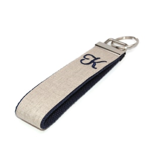 May include: A beige and navy blue key chain with a silver key ring. The key chain has the letter "K" embroidered on it in navy blue thread.