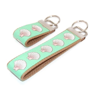 May include: Two keychains with a light green background and a pattern of white seashells with pink accents. The keychains are made of brown fabric and have silver key rings.