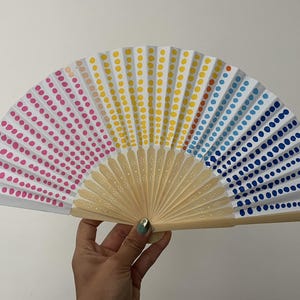 Peut inclure: Éventail à main avec un motif à pois colorés sur fond blanc. Le motif est divisé en sections de pois roses, jaunes et bleus. Le manche est en bois clair.