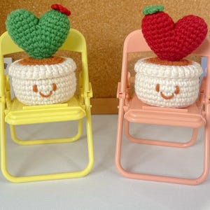Puede incluir: Dos plantas en miniatura de forma de corazón tejidas a crochet en macetas, cada una sentada en una pequeña silla plegable amarilla y rosa. Las plantas tienen caras sonrientes.