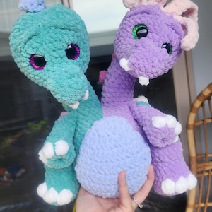 Könnte beinhalten: Zwei handgefertigte Häkeldrachen-Plüschtiere. Einer ist türkis, der andere lila. Beide haben große, glitzernde Augen und weiße Akzente. Sie sind durch einen hellblauen, eiförmigen Körper verbunden. Die Plüschtiere sind weich und kuschelig.
