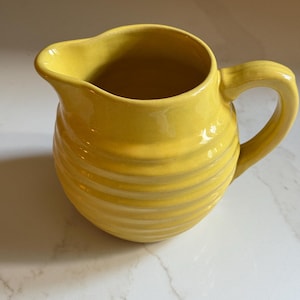 May include: A bright yellow ceramic pitcher featuring a rounded, bulbous body with horizontal ribbed detailing, a wide pouring spout, and a curved handle attached to the side. The surface has a smooth, glossy finish that reflects light.