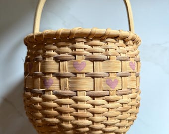 This is a charming handmade woven splint wood basket featuring an arched fixed handle and a sweet pattern of small painted pink hearts