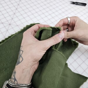 May include: Close-up of a person's hands sewing two pieces of dark green fabric together. The fabric is folded over and pinned in place.