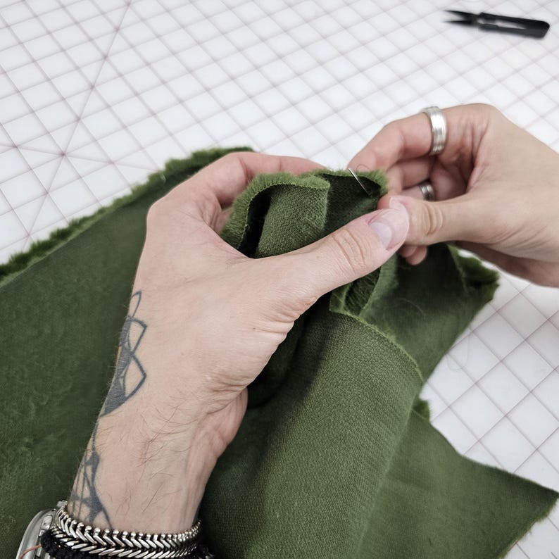 May include: Close-up of a person's hands sewing two pieces of dark green fabric together. The fabric is folded over and pinned in place.