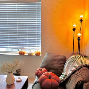 May include: A brown couch with a green and white throw blanket and two orange knitted pumpkin pillows. A floor lamp with three lights is in the background.