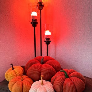 May include: Five decorative pumpkins in various colors and textures, including orange, white, and red, are arranged on a dark surface. The pumpkins are illuminated by a three-tiered candle holder with three lit candles.