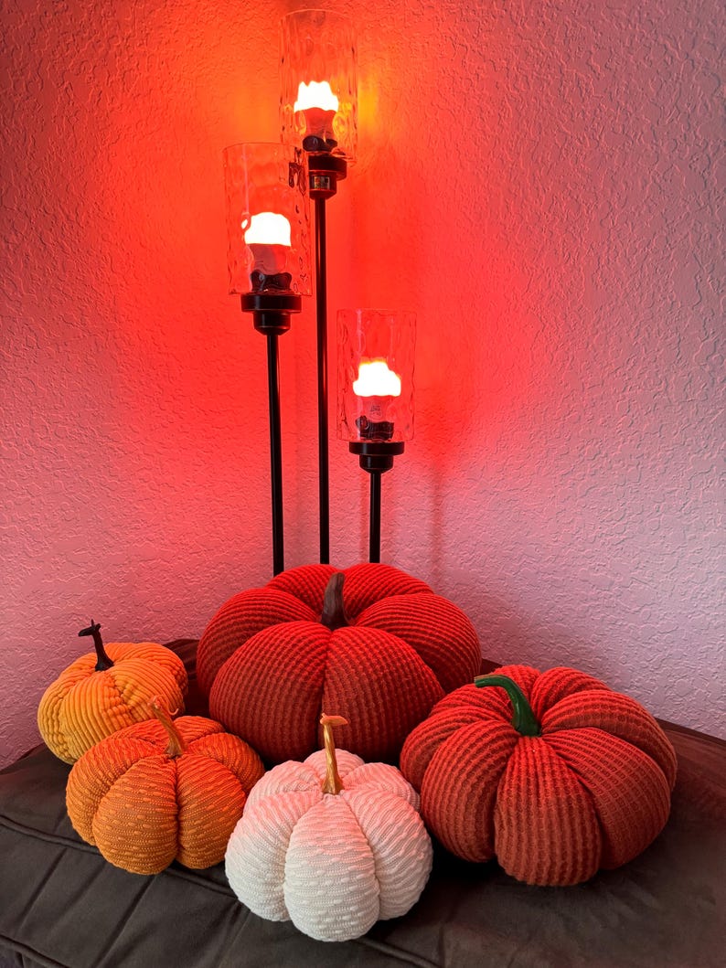 May include: Five decorative pumpkins in various colors and textures, including orange, white, and red, are arranged on a dark surface. The pumpkins are illuminated by a three-tiered candle holder with three lit candles.
