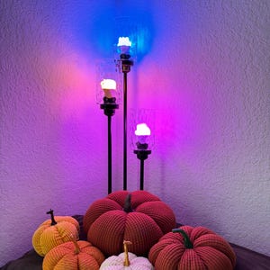 May include: Five corduroy pumpkins in various colors, including red, orange, and white, are arranged on a brown surface. Three black metal candle holders with glass shades are lit with colorful LED lights in the background.