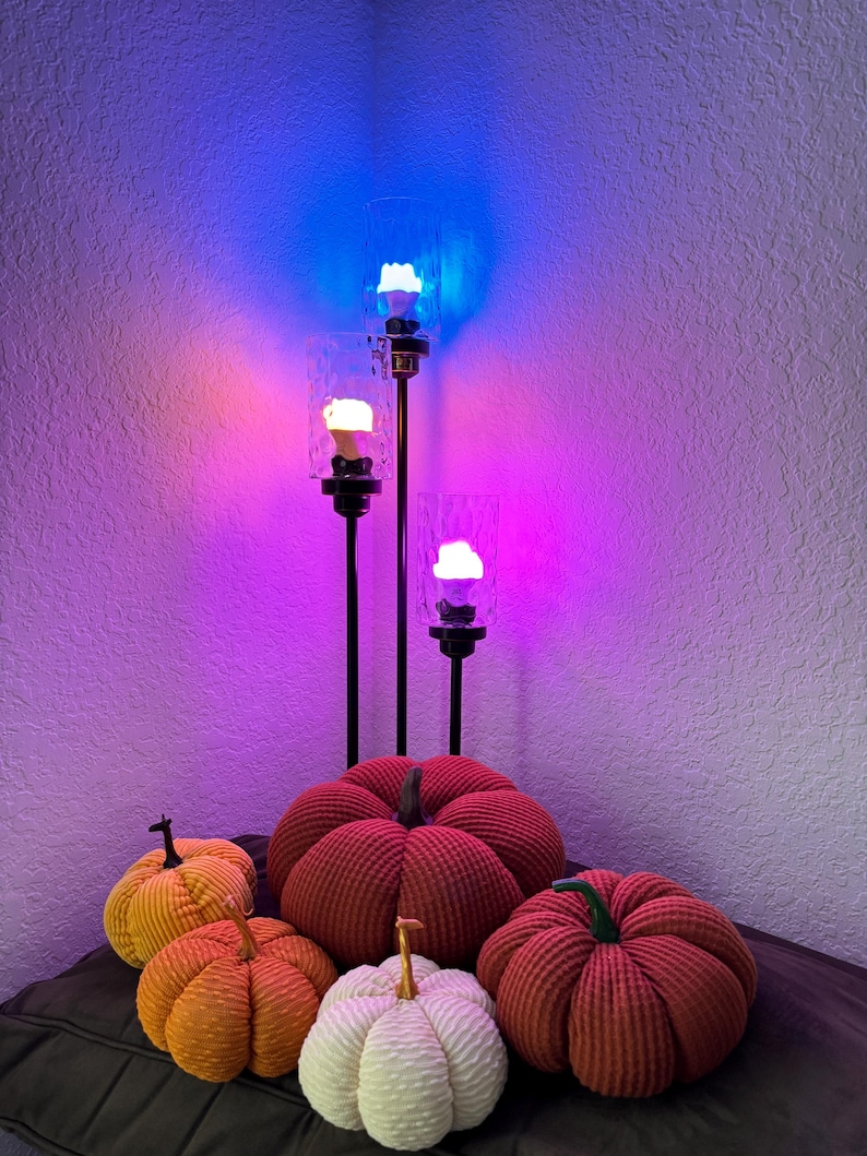 May include: Five corduroy pumpkins in various colors, including red, orange, and white, are arranged on a brown surface. Three black metal candle holders with glass shades are lit with colorful LED lights in the background.